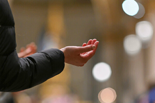 Person wearing a black jacket extends hands, palms open and facing up, in a reverent gesture inside a blurred warm interior.